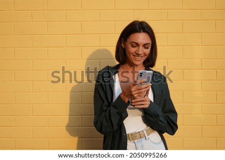 Caucasian woman wearing shirt at sunset on yellow brick background outdoor positive look at mobile phone screen with smile