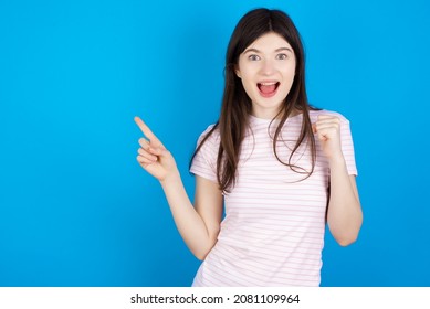 Caucasian woman wearing striped T-shirt over blue background points at empty space holding fist up, winner gesture. - Shutterstock ID 2081109964