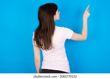 Caucasian woman wearing striped T-shirt over blue background pointing to object on copy space, rear view. Turn your back