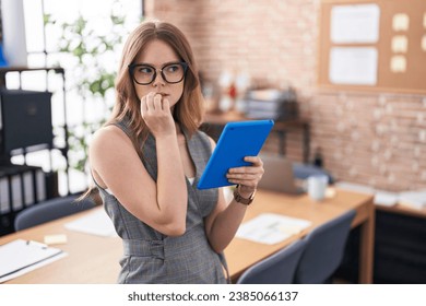 Caucasian woman working at the office wearing glasses looking stressed and nervous with hands on mouth biting nails. anxiety problem. 
