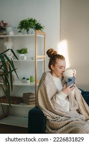 Caucasian woman wrapped in warm cozy plaid holding mug of tea, enjoying leisure at home while chatting with someone on mobile phone. Domestic lifestyle - Shutterstock ID 2226831829
