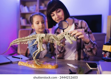 Caucasian women wearing casual clothes making prehistorical research. Happy little daughter and her mother sitting together at table and study fossil animals with magnifying glass at evening time. Two - Powered by Shutterstock