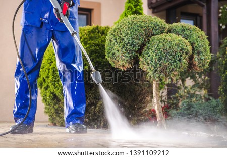 Caucasian Worker in His 30s with Pressure Washer Cleaning Residential Driveway. Garden and Home Surrounding Maintenance.