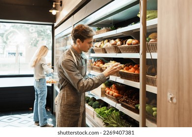 Caucasian young adult employee with an apron is looking at fruit on the shelves and checking the quality and freshness. Salesman with responsibility to quality check stock of fruit in grocery store.  - Powered by Shutterstock