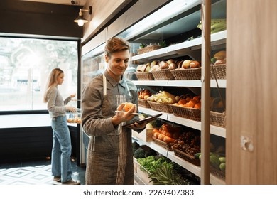 Caucasian young adult employee with an apron is checking quality of fruit on the shelves. Salesman is holding a tablet to record information about the quality and freshness of fruit in grocery store. - Powered by Shutterstock