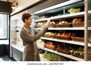 Caucasian young adult employee is holding corn and taking a picture with his smartphone to send to stock manager for quality checking. Salesman is checking the stock of fruit before opening the store. - Powered by Shutterstock