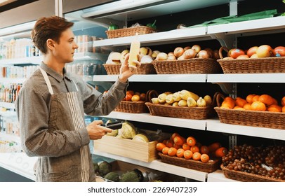 Caucasian young adult employee is working on checking the stock of fruit on shelves with barcode scanner. Salesman is holding a barcode scanner to examine the stock of corns in the basket on shelf. - Powered by Shutterstock