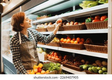 Caucasian young adult employee is working on adding the stock of fruit to the shelf before the grocery store opens. Active saleswoman with an apron is holding fruit basket and adding apple to shelf. - Powered by Shutterstock