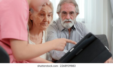 Caucasian young adult nurse is visiting elderly couple and holding a tablet to show medical records. Female caregiver is explaining result of medical examination and treatment to senior grandparents.  - Powered by Shutterstock