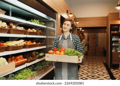 Caucasian young adult saleswoman is carrying a fruit basket and preparing to add stock of fruit to the shelf. Female employee who working in the grocery store looking at the camera with smile face.  - Powered by Shutterstock