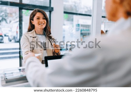 A Caucasian young adult woman is at the counter in the pharmacy purchasing medication from a pharmacist.
