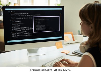 Caucasian young adult woman developing neural network code on desktop computer, sitting at desk, focusing on programming interface with lines of code visible on large monitor