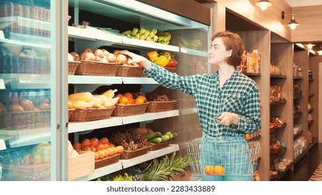 Caucasian young adult woman is picking up oranges and apples from the shelf and putting them in shopping basket for checkout. Female customer is shopping fruit for good health at the grocery store. - Powered by Shutterstock