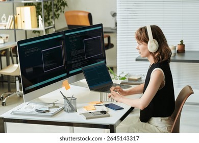 Caucasian young adult woman wearing headphones developing neural network code on desktop computer and laptop, sitting at desk with multiple monitors displaying programming interface