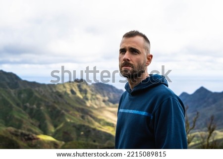 Caucasian young attractive man with blue hoodie and cloudy day, looking serious and observing the landscape