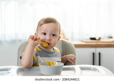 Caucasian Young Baby Toddler Eating Healthy Foods In Kitchen At Home. Little Adorable Cute Kid Boy Infant Sitting On Children Chair Alone, Enjoy Having Lunch In House. Child Development Skill Concept.