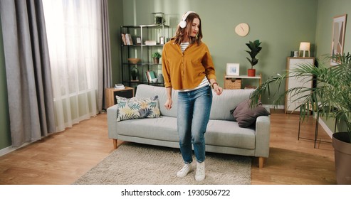 Caucasian young beautiful joyful woman dancing in modern living room in house alone listening to music in white headphones making moves and having fun. Rhythm concept, home dancer