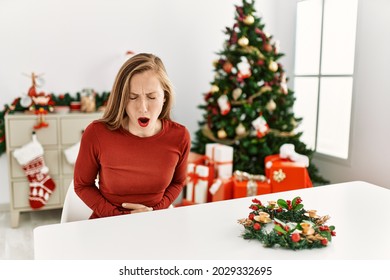 Caucasian Young Blonde Woman Sitting On The Table By Christmas Tree With Hand On Stomach Because Indigestion, Painful Illness Feeling Unwell. Ache Concept. 