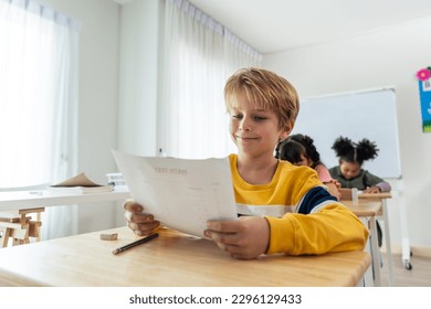 Caucasian young boy student doing an exam test at elementary school. Adorable children sitting indoors on table, feeling confidence while looking at new grade and learning with teacher at kindergarten - Powered by Shutterstock