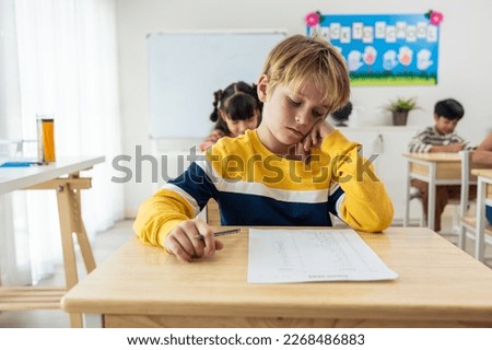 Caucasian young boy student doing an exam test at elementary school. Adorable children sitting indoors on table, feeling upset and depressed while writing notes, learning with teacher at kindergarten.