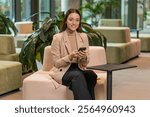 Caucasian young businesswoman girl using mobile phone texting share messages content working on smartphone social media applications online, relaxing in modern office lobby. Lady sitting in hotel hall