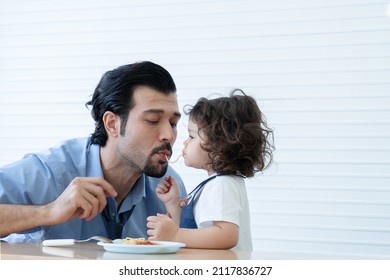 Caucasian Young Father With Beard And Little Cute Daughter Trying To Eat Spaghetti Same String At Home. Adorable Kid Girl Enjoy Eating With Face Is Mess Up With Tomato Sauce