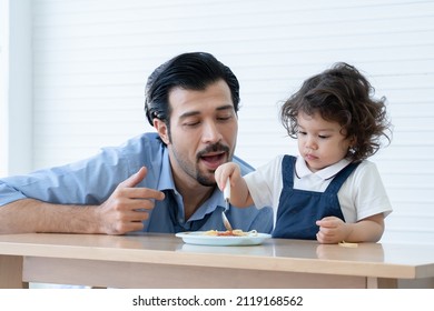 Caucasian Young Father With Beard Looking And Cheer Up Little Cute Daughter Trying To Eat Spaghetti With Spoon By Herself At Home And Adorable Kid Girl Enjoy Eating With Face Is Mess Up With Ketchup