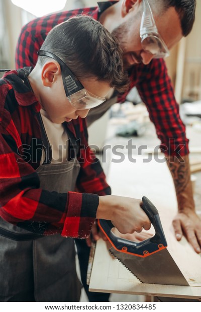 Caucasian young father in protective eyewear\
teaching his attentive pensive junior school son to saw wood in\
wooden workshop