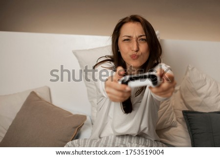 Caucasian young female with dark hair in white pajamas in bedroom with stylish lighting plays video games on the console using the joystick, smile and make antics. Quarantine entertainment concept.
