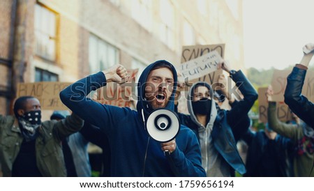 Caucasian young handsome guy protesting in middle of multiethnic crowd of protesters and screaming mottos in megaphone. Guy leading at manifestation for human rights.