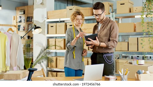 Caucasian Young Handsome Male Stylish Boss Tapping On Tablet Device Speaking With Female Designer Discussing New Clothing Collection, Clothing Shop, Warehouse, Delivery Store, Fashion Industry