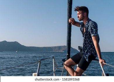 Caucasian young handsome man sitting on the rails of a sailboat in the mediterranean sea in Greece. Looking in the distance thoughtful wearing marine blue shirt and shorts. Place to add text.