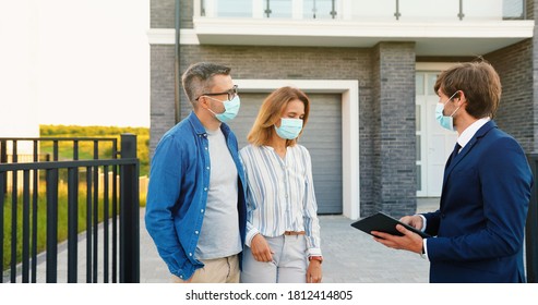 Caucasian young happy couple in medical masks buying house at outskirt and talking with male real-estate agent holding tablet device in hands. Outdoor. Coronavirus pandemic concept. - Shutterstock ID 1812414805