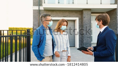 Caucasian young happy couple in medical masks buying house at outskirt and talking with male real-estate agent holding tablet device in hands. Outdoor. Coronavirus pandemic concept.