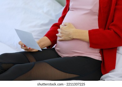 Caucasian young happy healthy female prenatal pregnant mother in casual outfit sitting smiling on bed in bedroom showing ultrasound Xray picture to family via video call with smartphone.