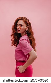 Caucasian Young Lady In Half Turn Looks At Camera With Impressive Gaze On Pink Background. Girl With Red Wavy Hair Holds Hands In Pockets Wearing Round Glasses And Casual Clothes. Beauty Concept.