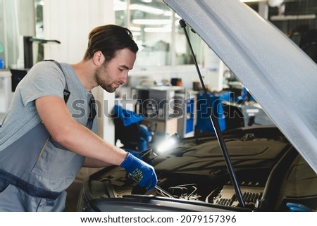 Caucasian young male car technician mechanic in special blue robe uniform holding pipe monkey wrench and flash light repairing fixing car at vehicle service inspection. MOT