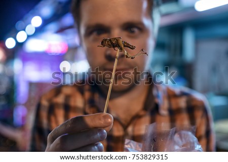 Caucasian young male eating cricket at night market in Thailand. Eating insect concept