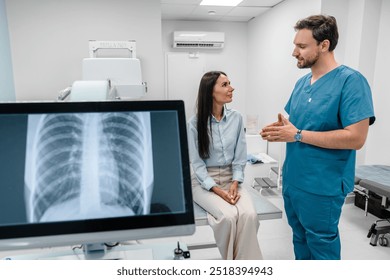 Caucasian young male medic doctor specialist radiologist in blue uniform explaining diagnosis on lung x-ray fluorography to female patient during medical appointment in hospital. - Powered by Shutterstock