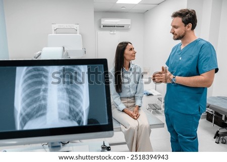 Caucasian young male medic doctor specialist radiologist in blue uniform explaining diagnosis on lung x-ray fluorography to female patient during medical appointment in hospital.