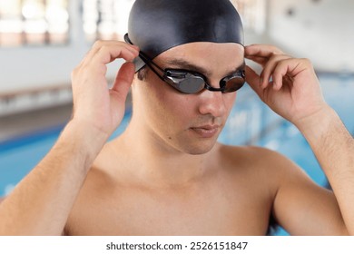 Caucasian young male swimmer adjusting goggles indoors, standing by pool. He has short dark hair, wearing a black swim cap and goggles, unaltered. - Powered by Shutterstock