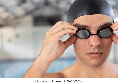 Caucasian young male swimmer adjusting goggles indoors, standing by pool, copy space. He has short brown hair, fair skin, and is wearing a black swim cap - Powered by Shutterstock