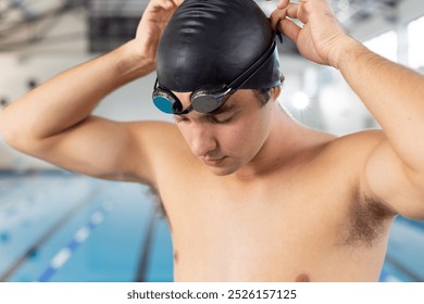 Caucasian young male swimmer adjusting swim cap, standing poolside indoors. He has short brown hair, light skin, and is wearing dark goggles. - Powered by Shutterstock