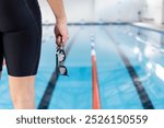 Caucasian young male swimmer holding goggles stands by the pool indoors, copy space. He has short brown hair, wearing a dark swimsuit, ready for training, unaltered