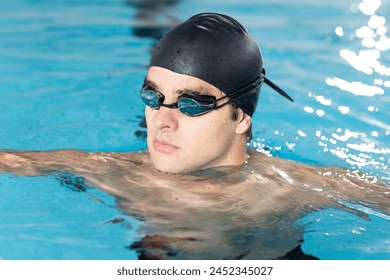 Caucasian young male swimmer resting indoors in a pool, wearing a black cap and goggles. He has short hair, fair skin, and is looking away from the camera, unaltered. - Powered by Shutterstock