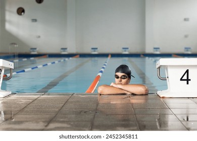 Caucasian young male swimmer resting at poolside indoors, wearing goggles, copy space. He has short dark hair, fair skin, and is in a swimming cap, unaltered - Powered by Shutterstock