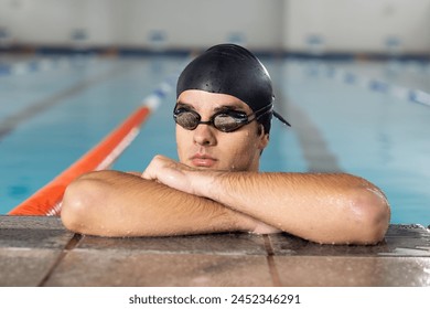 Caucasian young male swimmer resting at pool edge indoors, looking forward. He has short hair under a black cap, wearing goggles, unaltered. - Powered by Shutterstock