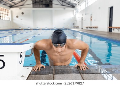 Caucasian young male swimmer resting at swimming pool edge indoors, wearing goggles. Blue background enhancing his short brown hair, fit body, and focused expression, unaltered - Powered by Shutterstock