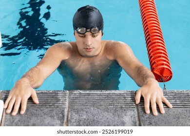 Caucasian young male swimmer resting at swimming pool edge indoors, looking focused. Wearing goggles and swim cap, holding onto edge, preparing for another lap, unaltered - Powered by Shutterstock