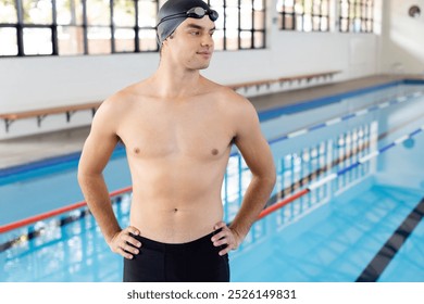 Caucasian young male swimmer standing by poolside indoors, looking confident. Wearing black swim cap and goggles, showcasing fit physique, unaltered. - Powered by Shutterstock